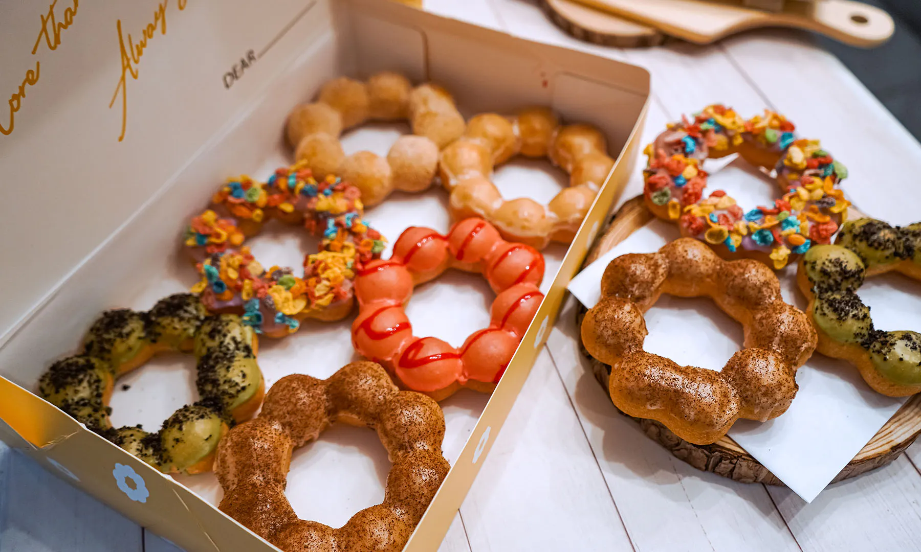Assortment of colorful ring‑shaped mochi donuts inside open paper box and on wooden platters, with matcha glaze, fruity cereal toppings, cinnamon dust and red syrup drizzle on white wooden table.