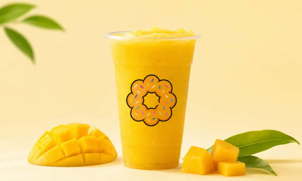 Bright yellow mango slushie in branded clear plastic cup with flower‑shaped logo. Sliced fresh mango chunks and green mango leaves arranged beside cup, warm pale yellow background.