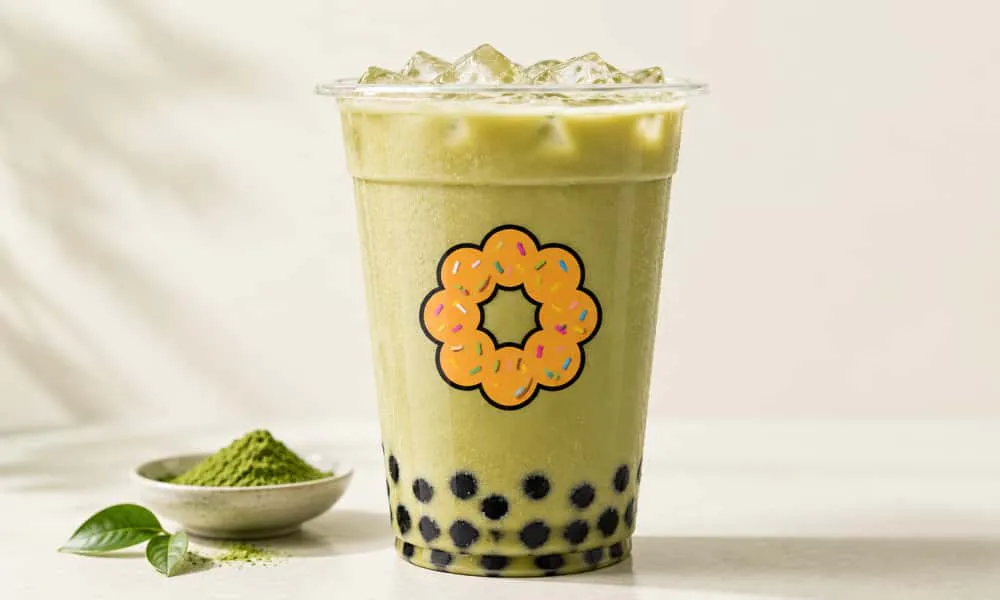 Creamy light‑green iced matcha bubble tea with black tapioca pearls at bottom of branded plastic cup. Small bowl of matcha powder and green tea leaves sit on light‑textured countertop.