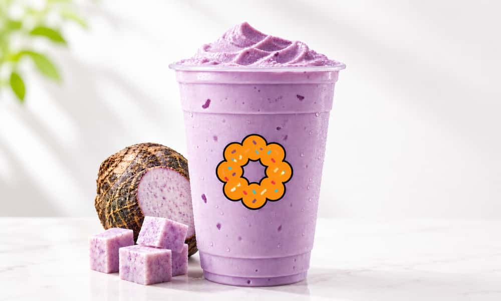 Lavender‑purple creamy taro slush piled high on branded cup. Whole taro root and cubed taro pieces next to cup, soft blurred green foliage in light‑toned background.