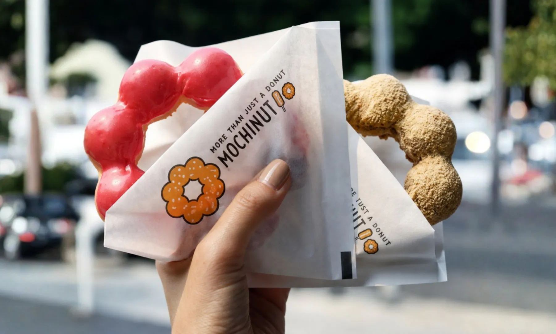 Hand holding wrapped pink and brown mochi donuts outdoors